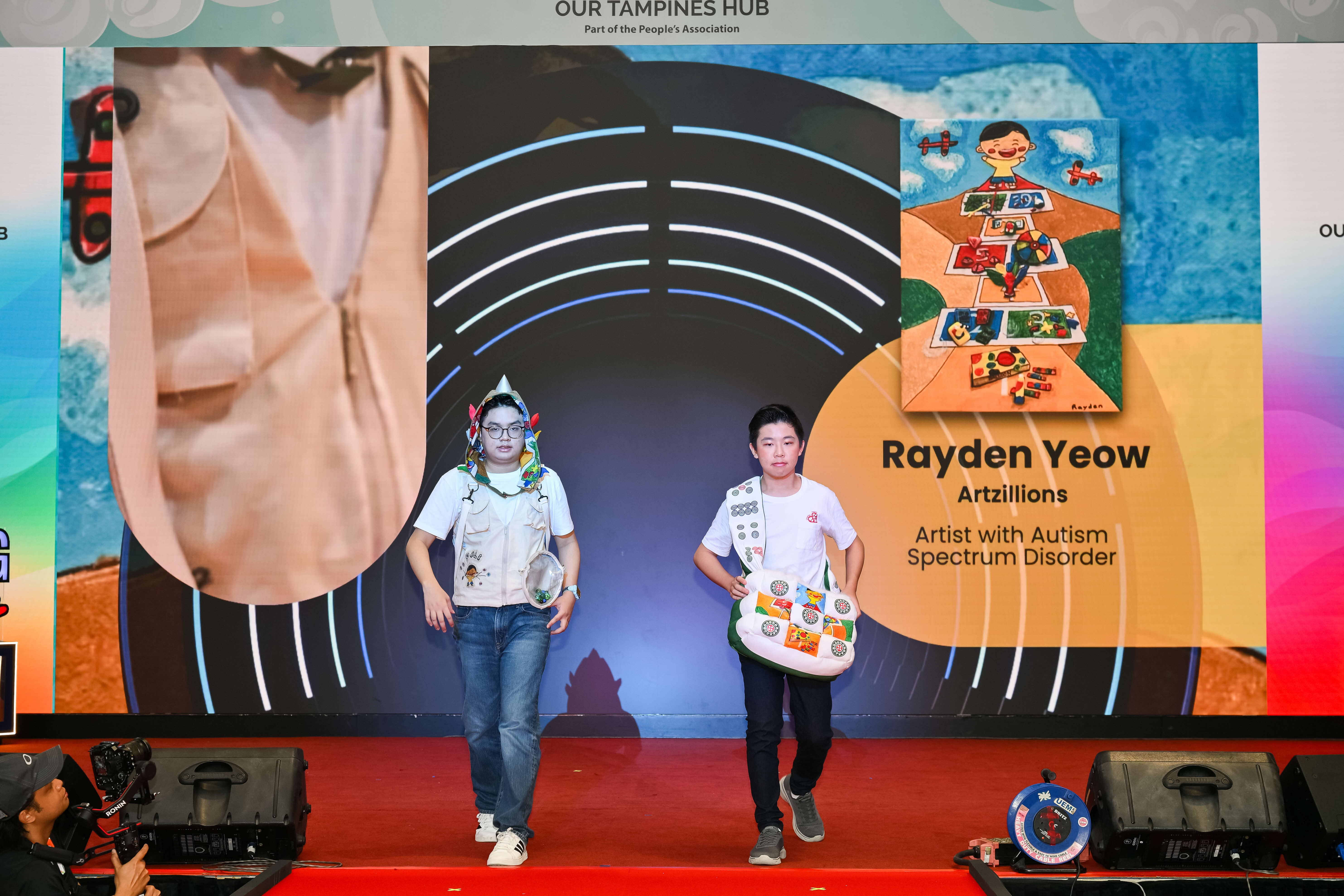 Two youths walk across a stage during an event, with a large screen behind them displaying artwork and text